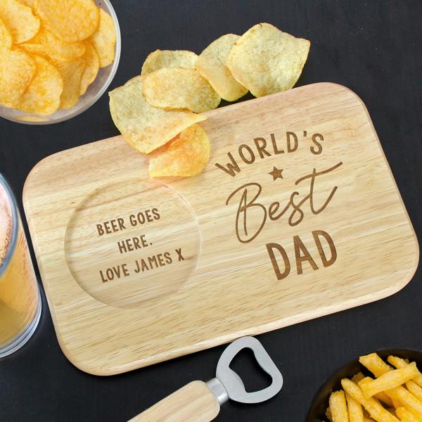 A rectangular wooden coaster tray on a black surface featuring an engraved "World's Best Dad" design, a circular groove holding a glass of beer, and a handful of crisps on the side, next to a bottle opener.