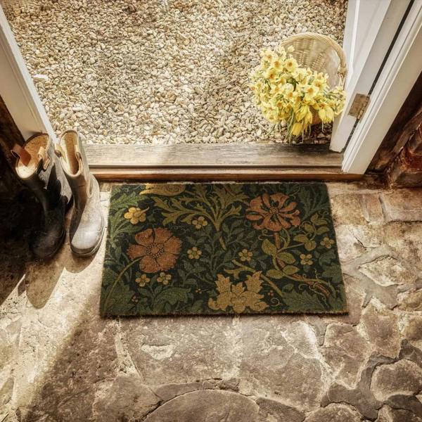 Decorative doormat with floral pattern on stone floor, next to shoes and a basket of flowers.