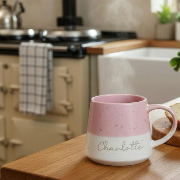 A pink and white two-tone speckled ceramic mug resting on a wooden kitchen counter, with steam rising from the mug and the name "Charlotte" engraved on the side.