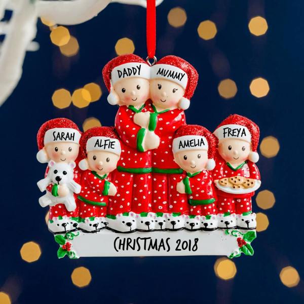 A colourful resin family Christmas decoration featuring six characters in matching red polka dot pyjamas and glittery Santa hats, handwritten with names like Daddy, Mummy, Sarah, Alfie, Amelia, and Freya, reading 'Christmas 2018' against a dark blue background with bokeh fairy lights.