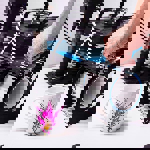 White ceramic mug with pink floral design next to a mug heat press machine and a person's hand adjusting it.