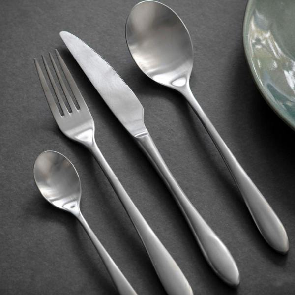 A 4-piece place setting of brushed silver stainless steel cutlery including a teaspoon, dinner fork, serrated knife, and dessert spoon resting on a dark slate surface beside a ceramic plate.