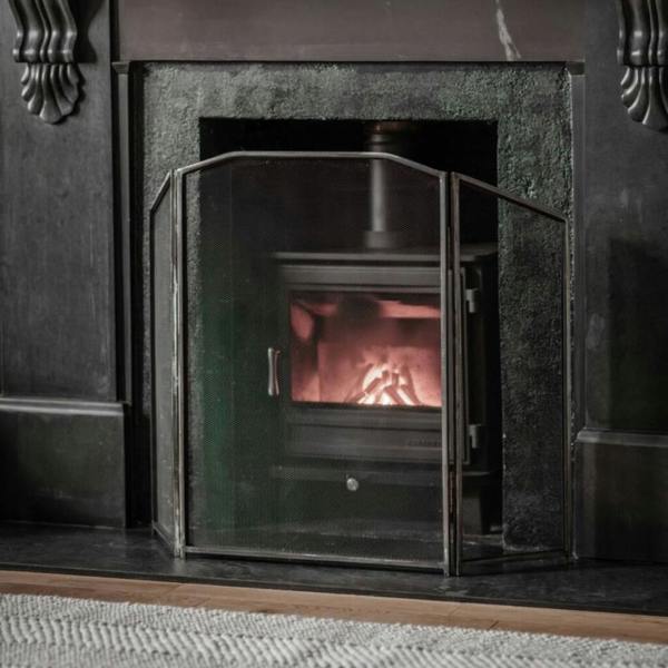 A close-up view of a robust, three-panel folding antique pewter firescreen, showing its decorative arched top, fine mesh panels, and solid frame construction as it stands before a glowing fireplace.