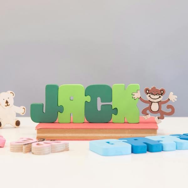 A colourful wooden name puzzle spelling the name 'JACK' in interlocking green letter pieces. The jigsaw stands on a pink book and is accompanied by small wooden decorative pieces, including a white bear and a brown monkey.
