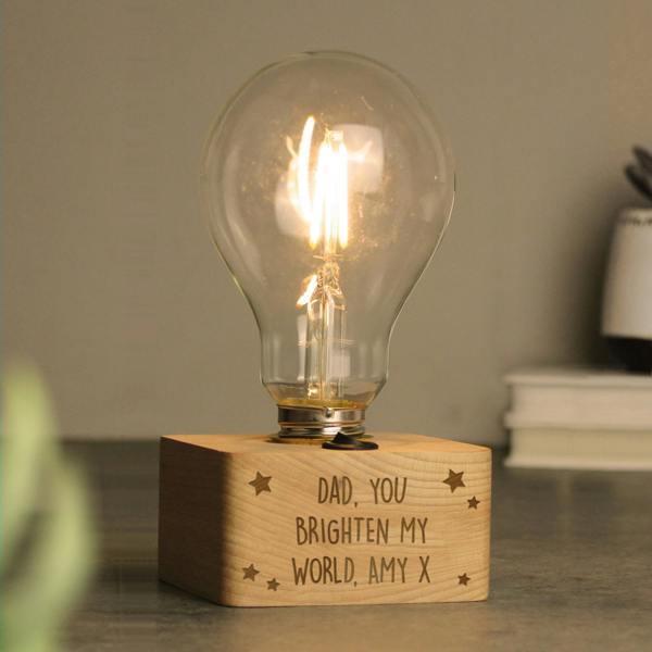 A personalised stars LED bulb table lamp by Abode Home & Gifts with a solid wooden base engraved with "DAD, YOU BRIGHTEN MY WORLD, AMY X", styled with a stack of books and a small potted plant in the background.