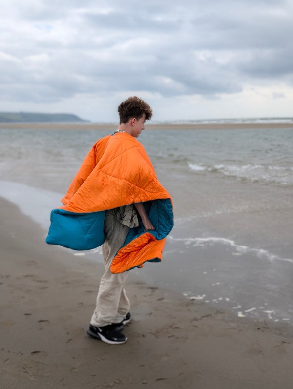 A man stood on the beach watching the waves, wearing a blue/Orange reversible Blanko as a poncho with the orange side visible.