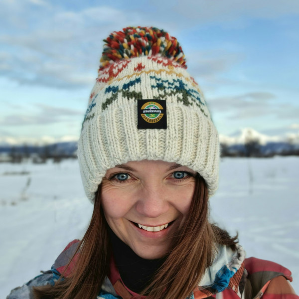 Woman in the snowy foothills of Northern Norway is wearing a chunky Nordic knit bobble hat. The oatmeal base and warm autumn toned Nordic design hat also has a deep ribbed headband, contains reflective yarns and has a luxury sherpa fleece lining.