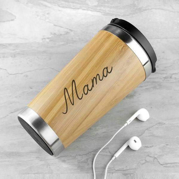 A personalised bamboo travel mug engraved with the name 'Mama' lying diagonally on a grey textured surface next to white wired earphones.