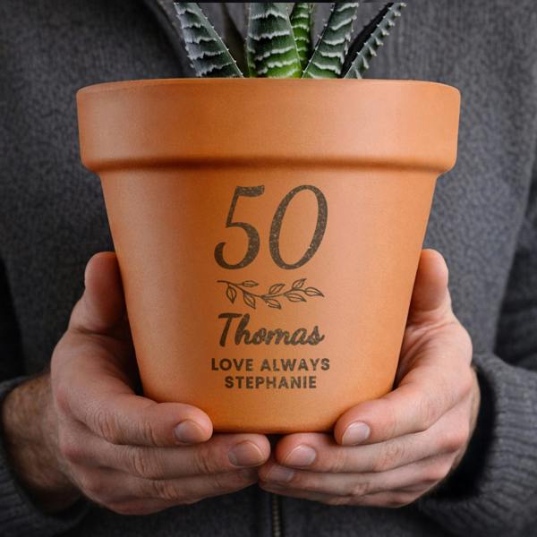 A person holding a natural terracotta plant pot engraved with the age 50, a leaf motif, the name Thomas, and the message LOVE ALWAYS STEPHANIE.