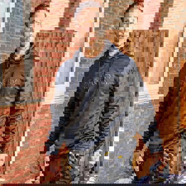 Stanley Hudson Mens Applique Logo Work Hoodie, a man wearing a dark grey hoodie with the Stanley logo embroidered on the front, standing outdoors near a brick wall and wooden fence.