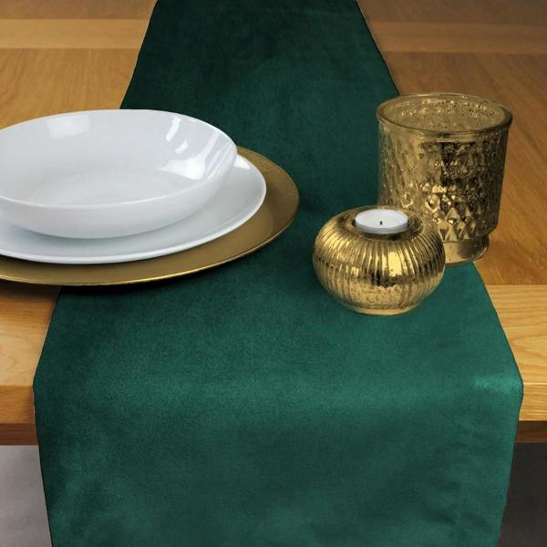 Luxury emerald green velvet table runner on a wooden dining table set for a formal meal with white bowls and gold chargers.