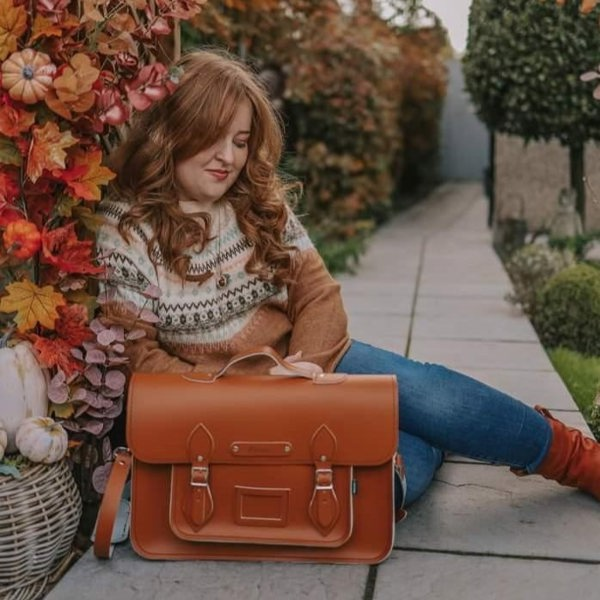 Our model, Emily, is sitting on the ground next to autumnal leaves, with a Burnt orange Zatchels satchel on the ground next to her. 