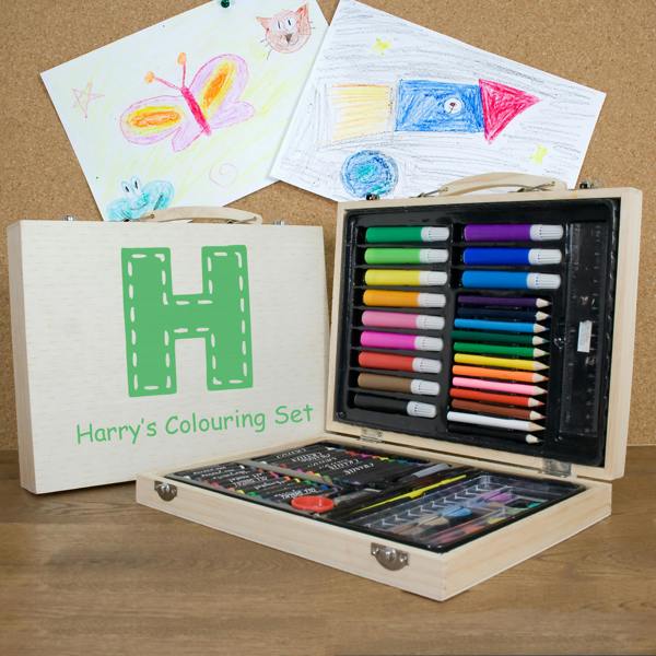A 63-piece personalised children's colouring set in a wooden case printed with a large green letter H and Harry's Colouring Set, open on a desk displaying art supplies alongside drawing artwork.