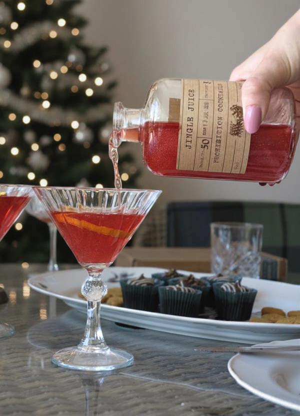 A hand with pink nail polish pours a red cocktail from a clear glass bottle labeled 'Jingle Juice' by 'Prohibition cocktails' into a martini glass on a table, with a blurred Christmas tree in the background.