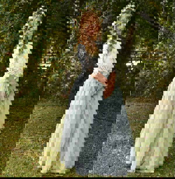 Young girl in a white lace top and light green tulle skirt standing outdoors with greenery in the background.