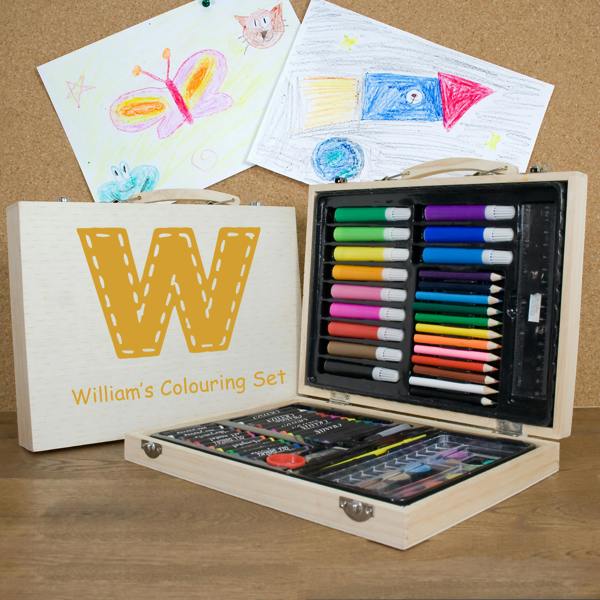 A 63-piece personalised children's colouring set in a wooden case printed with a large yellow letter W and William's Colouring Set, open on a desk displaying art supplies alongside drawing artwork.