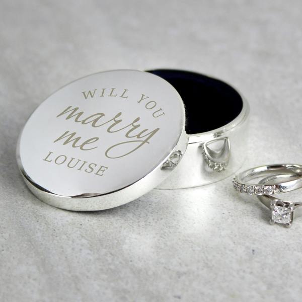 A polished silver round ring box with an open lid engraved with 'WILL YOU marry me LOUISE', styled on a light stone countertop next to a diamond engagement ring and wedding band.