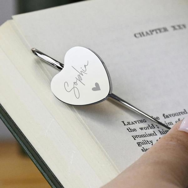 A polished silver-coloured heart bookmark engraved with the name Sophia in script text, marking a page inside an open book.