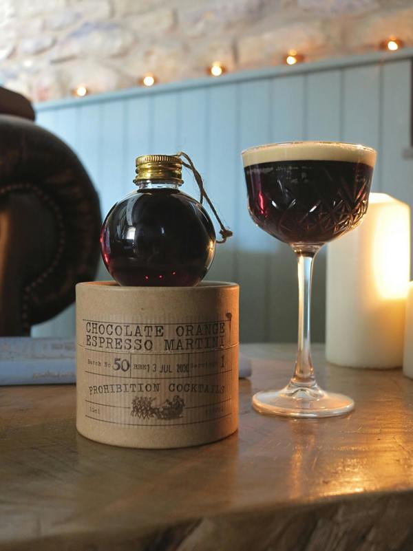 A small dark glass bottle with a gold cap sits on a cylindrical wooden box labeled 'Chocolate Orange Espresso Martini', next to a coupe glass filled with the dark cocktail, on a wooden table with warm candlelight in the background.