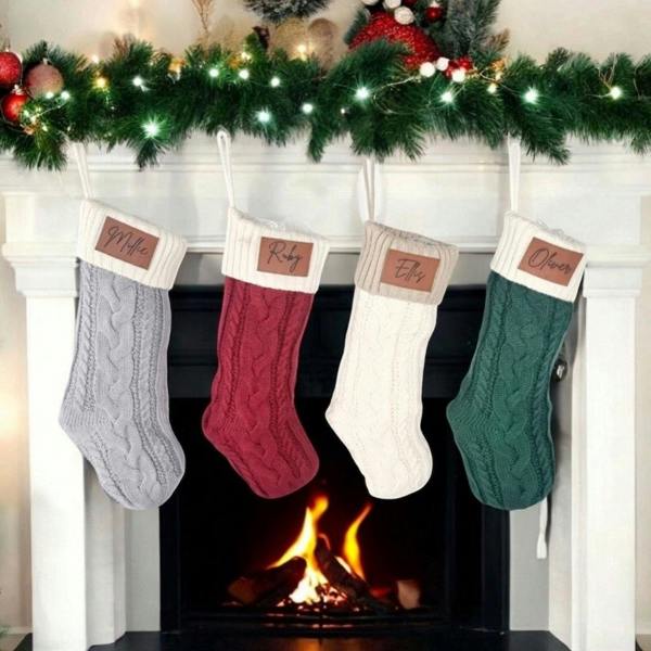 Four personalised cable-knit Christmas stockings in grey, red, cream, and forest green hanging from a white fireplace mantle with a glowing hearth.