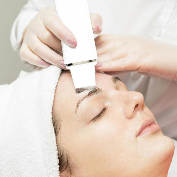 A person with a towel around their head receives a facial treatment as a gloved professional uses the Rio Ultrasonic Skin Cleanser on their forehead.
