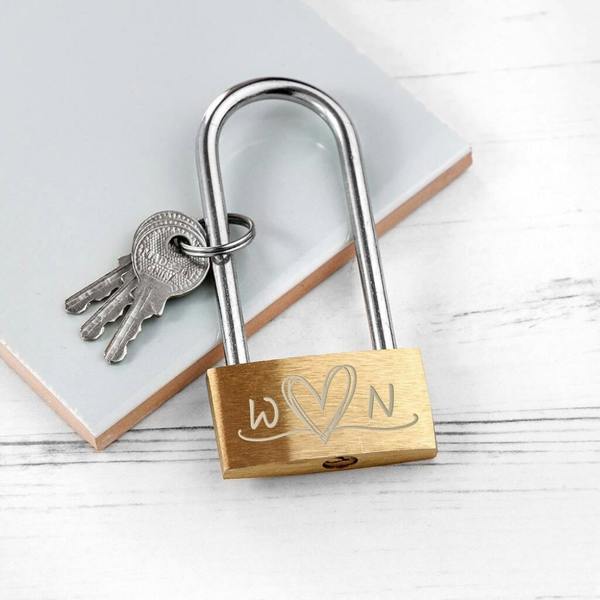 A solid brass love padlock engraved with the initials 'W' and 'N' connected by a heart, shown with three metal keys on a white wooden surface.