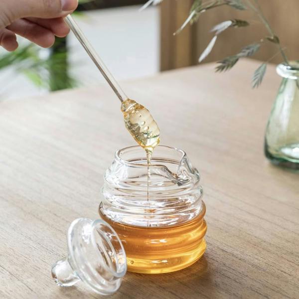 Clear hand-blown recycled glass honey pot filled with golden honey, featuring a matching glass honey dipper and lid on a wooden table.