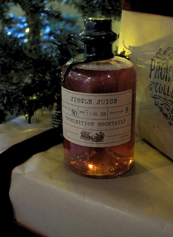 A small glass bottle filled with amber liquid, featuring a vintage-style cream label that reads 'JINGLE JUICE' and 'PROHIBITION COCKTAILS,' with a dark stopper. The bottle is photographed on a table with holiday greenery and warm ambient lighting in the background.