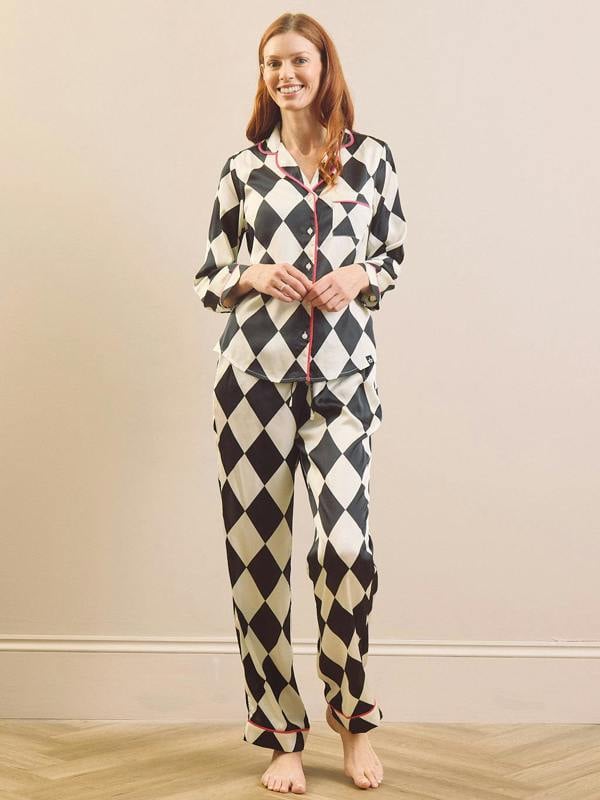 A woman modeling a two-piece pyjama set with a black and cream harlequin diamond pattern, featuring a button-front top with red piping and matching patterned pants, set against a neutral beige wall and light wood floor.