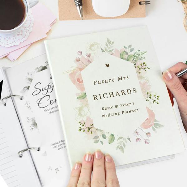 A top-down shot of a ring-bound wedding planner with a soft green cover and a watercolour floral frame, showing the custom-printed text 'Future Mrs RICHARDS Katie & Peter's Wedding Planner' held by two hands on a desk next to a coffee cup.