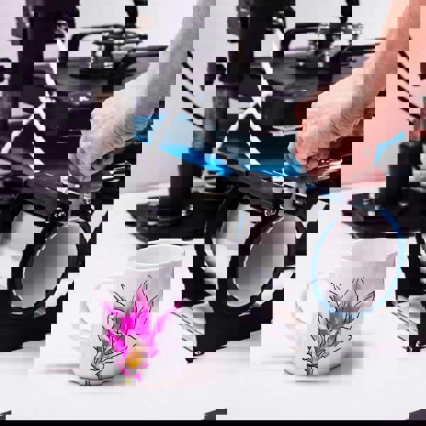 White ceramic mug with pink floral design next to a black and blue heat press machine on a white surface.