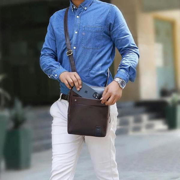 Man in blue shirt and white pants holding a brown leather messenger 
 bag outdoors.