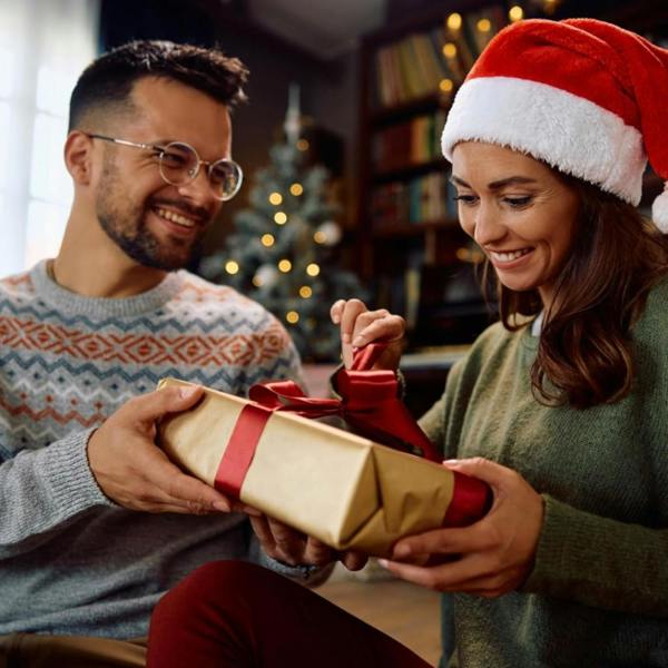 Man giving a gold gift-wrapped Christmas present with a red satin bow to a smiling woman wearing a Santa hat.