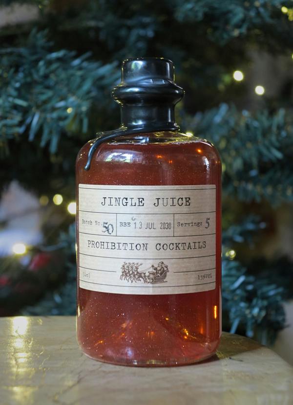 A vintage-style glass bottle filled with amber-red liquid, featuring a black stopper and a cream paper label that reads 'JINGLE JUICE' and 'PROHIBITION COCKTAILS.' The bottle is photographed on a tabletop with blurred evergreen branches and warm string lights in the background.