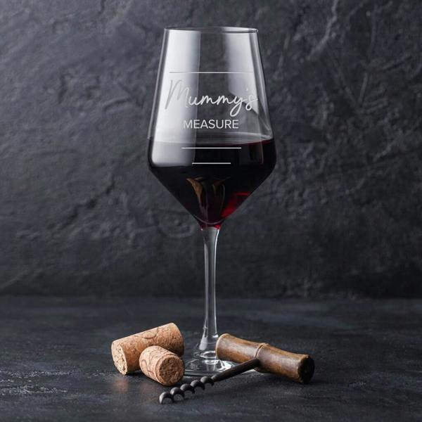 A clear stemmed wine glass filled with red wine, showing white text that reads Mummy's Measure next to three tiered lines.