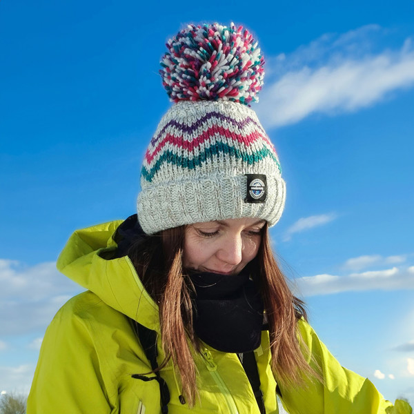 Women in a snowy landscape is wearing a chunky knit bobble hat with pink and teal peaks pattern on a white and pale grey base with a deep ribbed headband. The hat also contains reflective yarns and has a luxury sherpa fleece lining.