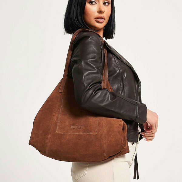 Image shows a woman carrying a tan suede bag over her shoulder. The bag features subtle embossing which reads: Barneys Originals.