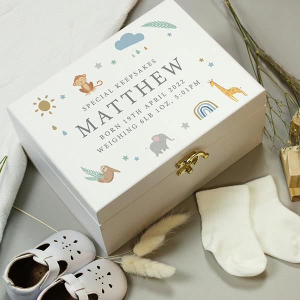 A rectangular hand-painted white wooden keepsake box with a gold metal clasp sits angled on a grey surface. The lid is printed with illustrated safari animals including an elephant, giraffe, monkey, and sloth around the capitalised custom birth text 'SPECIAL KEEPSAKES, MATTHEW, BORN 19TH APRIL 2022, WEIGHING 6LB 10OZ, 5:01PM'. White baby shoes and cream socks are arranged in the foreground.