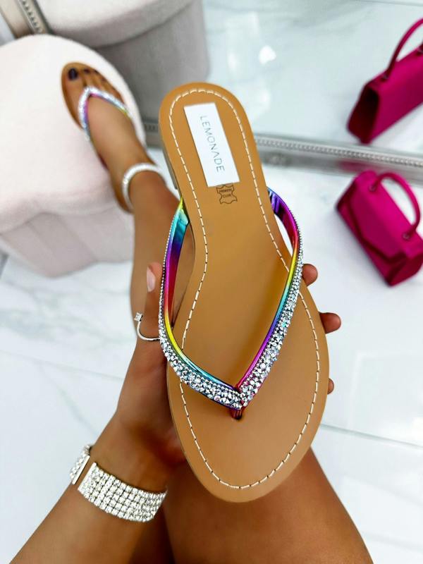 Multicolored sandal with rhinestone details held by a hand, with a blurred background of pink handbag and white shoes.