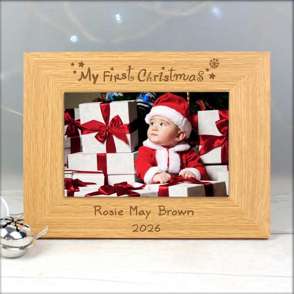 An oak veneer landscape photo frame engraved with 'My First Christmas' at the top and 'Rosie May Brown 2026' at the bottom, holding a picture of a baby in a Santa suit surrounded by gifts.