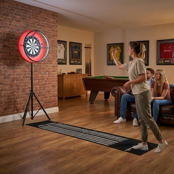 Dartboard LED ring light with vibrant red glow, mounted on a tripod, illuminating a player aiming at the dartboard in a cozy game room
