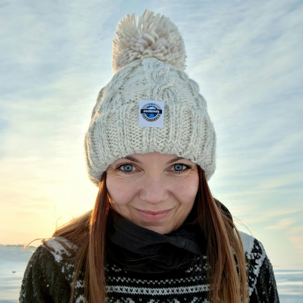 Woman in a snowy tree lined glade is wearing a cream oatmeal chunky cable knit Super Bobble hat. The hat also has a deep ribbed headband and contains reflective yarns and a luxury sherpa fleece lining.