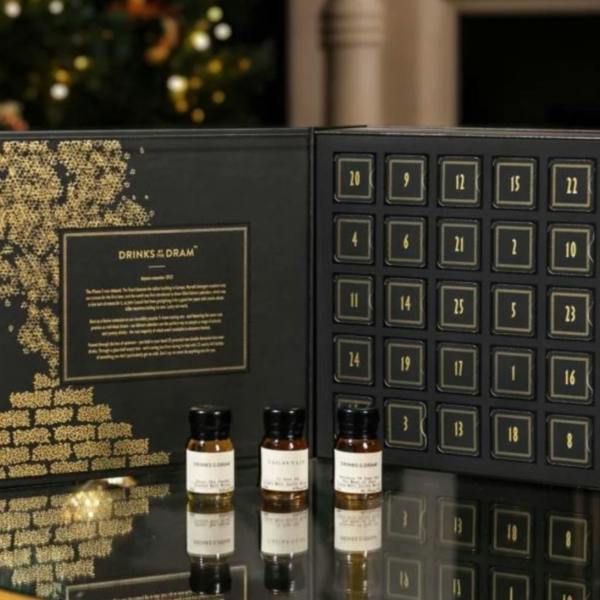 An open black and gold Drinks by the Dram Peaty Whisky Advent Calendar displaying numbered doors from 1 to 25 alongside three small wax-sealed dram bottles.