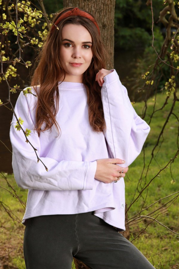 This is a young woman looking at the camera. She is standing in front of a tree which has yellow leaves. She has long hair with a red band. Her left hand is near her chin while her right hand is holding her left elbow. She is wearing a bella oversized cropped sweat shirt in lilac. Here trousers are grey.