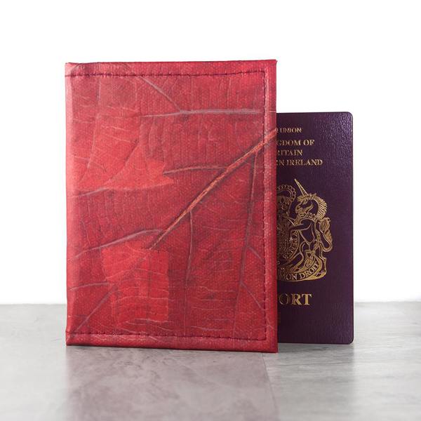 A red Leaf Leather Passport Cover made from dried, natural leaves, standing upright next to a UK passport on a light wooden surface.