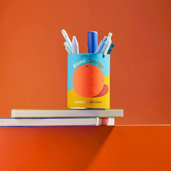 An empty blood orange drinking chocolate metal tin repurposed as a desk pencil holder, filled with blue and pastel pens and pencils on a stack of notebooks against a vibrant orange background.