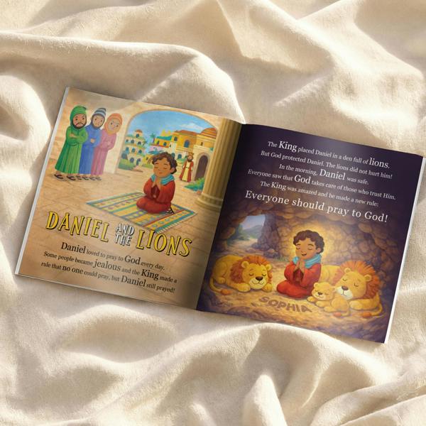 A flat lay product photograph showing an open page inside the Personalised Children's Bible Stories Book from Abode Home & Gifts, featuring a beautifully illustrated story titled 'Daniel and the Lions' with the name Sophia integrated into the cave floor illustration next to sleeping lions.