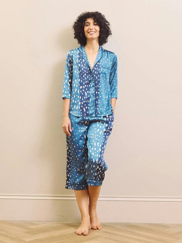 A woman modeling a two-piece capri pyjama set with a blue and white abstract pattern, standing barefoot against a beige wall and light wood floor.
