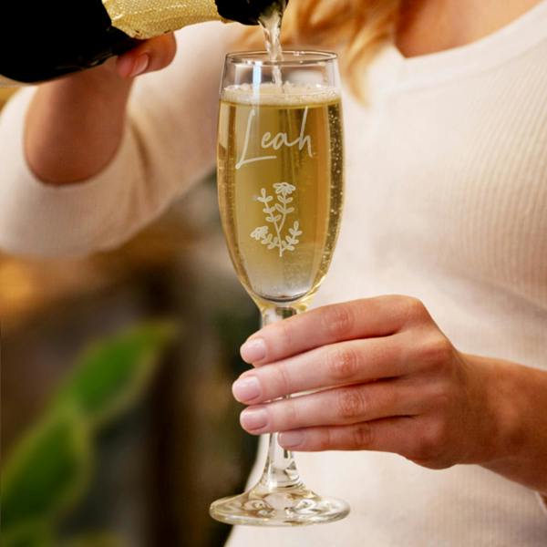 A close-up shot of a woman's hand holding a tall champagne flute by Abode Home & Gifts as it's being topped up with sparkling wine from a gold bottle. The glass is custom engraved with the name "Leah" in a script font above a delicate fine-line wildflower motif.
