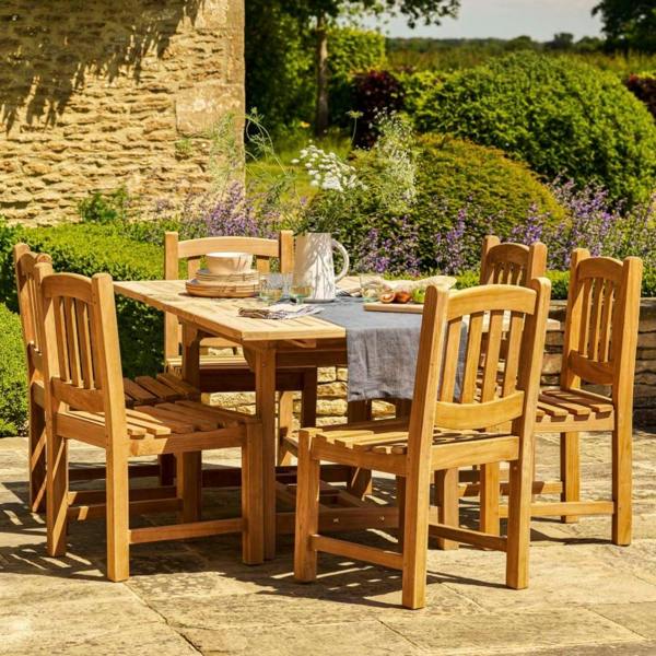 Rectangular solid teak extending dining table with six chairs on a stone patio, featuring a white flower vase and place settings.